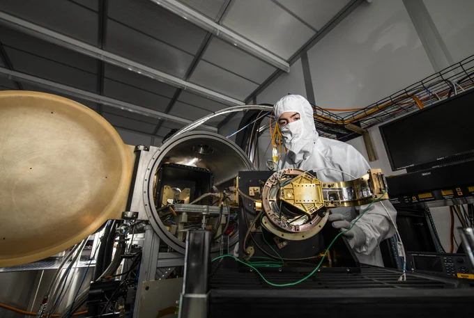 Scientist in cleanroom suit handling a complex gold-plated scientific instrument inside a high-tech laboratory.