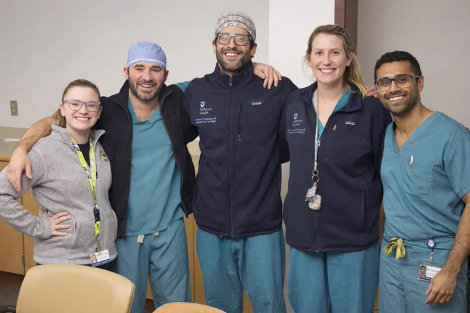 A group of five trauma team members standing close together in a clinical room. They are wearing teal scrubs or fleece jackets with Jefferson Hospital branding, and each person has an ID badge visible. The group is posed with arms around one another, suggesting a team photo. The background shows beige walls, cabinetry, and a table with chairs in the foreground.