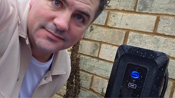 A selfie of a man with blue eyes next to an EV charging device branded \"INDRA\". The charging device is mounted on a brick wall.