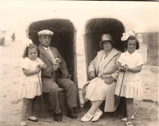 Vintage black and white photo showing four people sitting in front of beach huts. Two adults and two children each hold a small shovel and pose for the photograph.
