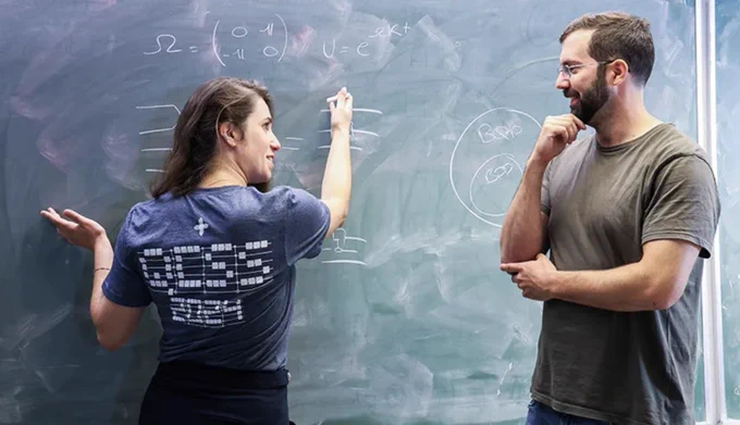 Two people discuss complex equations written on a large chalkboard in a classroom setting.