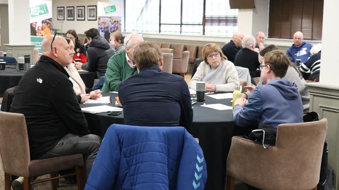 Seven people seated around a table, in discussion. Other tables and two Level Playing Field banners are in the background.