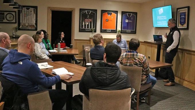 Level Playing Field's Daniel Townley is to the right of frame, presenting to a room of people, seated around three tables.