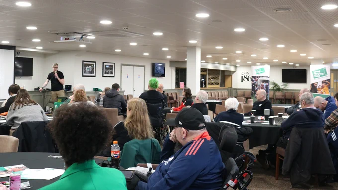 A wide shot from the right of a room, with people seated around tables. Level Playing Field head of fan engagement and supporter services, Liam Bird, is presenting at the front of the room.
