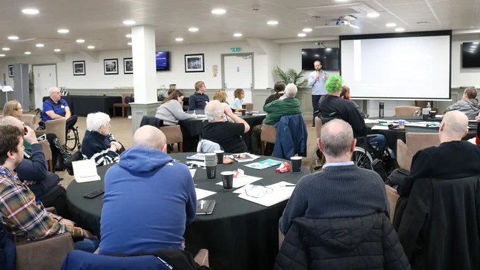 A wide shot from the right of a room, with people seated around tables. Level Playing Field chief executive, Owain Davies, is presenting at the front of the room, next to a projector screen.