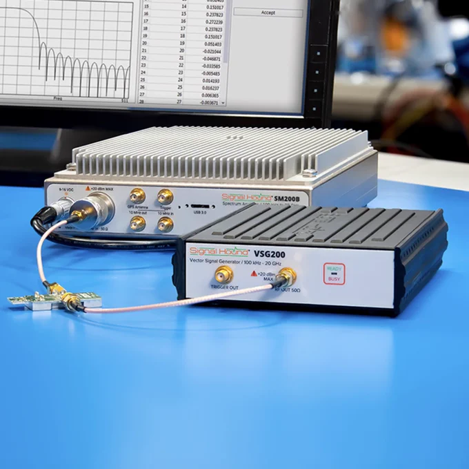 Signal Hound SM200B and Signal Hound VSG200 devices on a table, with cables connected. A monitor in the background displays a graph.