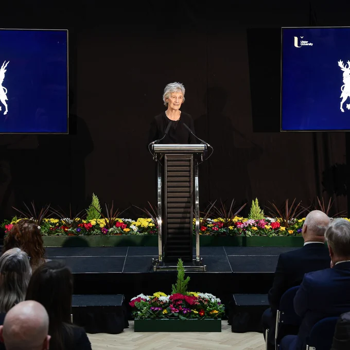President of Ireland Catherine Connolly stands at a podium delivering a speech to an audience seated in front of two large screens.
