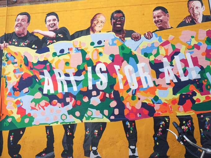 Group of people holding a colourful banner that reads 'ART IS FOR ALL' against a bright yellow background.