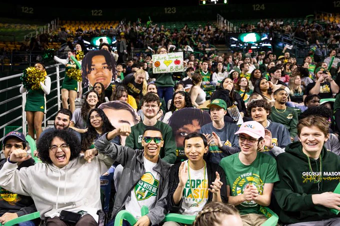 A large crowd of enthusiastic fans watches a game in an indoor arena. People in the front rows are cheering and wearing green and yellow team colors, some with George Mason apparel. On the left, a cheerleader holds gold and green pom-poms. The stands are packed, and some spectators are holding up cardboard cutouts of faces.