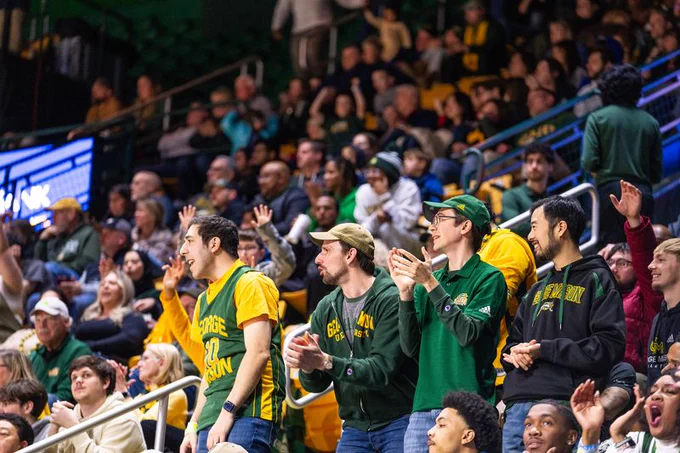 A group of male spectators wearing green and gold team apparel are cheering animatedly in the front row of a crowded indoor basketball arena. The fans are likely supporting the George Mason Patriots, whose official colors are green and gold.