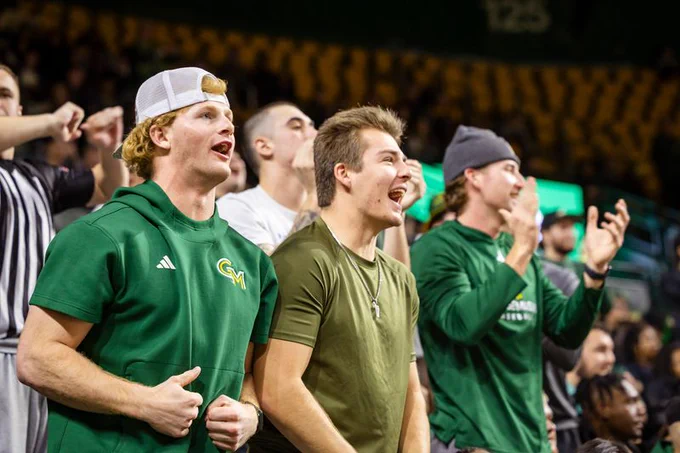 A close-up view of three young men excitedly watching an event in a crowded arena. The man on the left wears a green hoodie with a white baseball cap and a "GM" logo. The man in the center wears an olive green T-shirt with a cross necklace, and the man on the right wears a dark green long-sleeved shirt and a dark grey beanie. All three are animated and appear to be cheering for the same team.