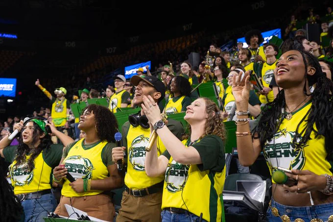 Members of the George Mason University "Green Machine" pep band are energetically playing instruments and cheering in the stands of a basketball game. Several individuals, wearing lime green and dark green tops with the words "GREEN MACHINE" and "NECHING" (likely part of the word "MECH NECHING" seen on a nearby sign), are featured in the foreground of the indoor arena.