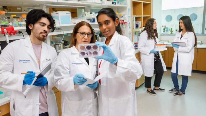 A group of individuals in lab coats working in a laboratory at UT Southwestern. Three people are examining a tray with samples, while two others are engaged in a discussion in the background.