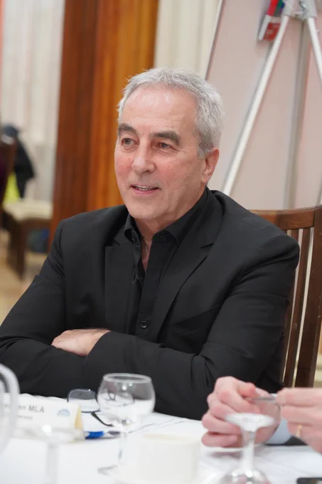 Deputy Chair Pat Sheehan, wearing a dark suit and dark shirt, sitting at a table with his arms crossed. In the foreground there are water glasses and a name place card.