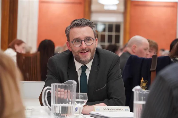 Nick Mathison leaning over a table wearing a dark suit and tie, with white shirt. In the foreground there is a water jug and a glass on a table. In the background other tables with discussions ongoing and orange coloured walls of the Long Gallery at Parliament Buildings