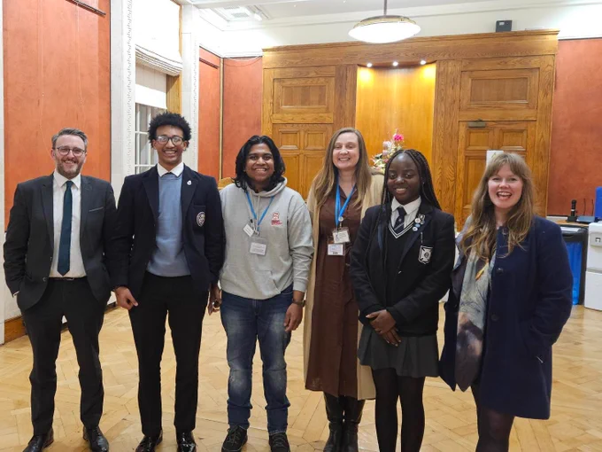 Group image showing six people, two of them wearing school uniforms, in the Long Gallery. In the background there are the orange walls of the room and a wooden door frame. To the far left of the image is the Committee Chair, Nick Mathison, wearing a dark suit and white shirt.