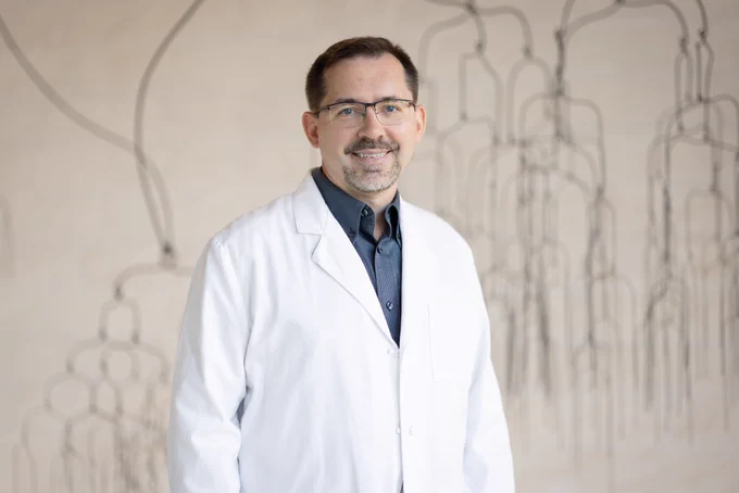 A person with a white lab coat and dark blue shirt standing in front of a beige wall with line art.