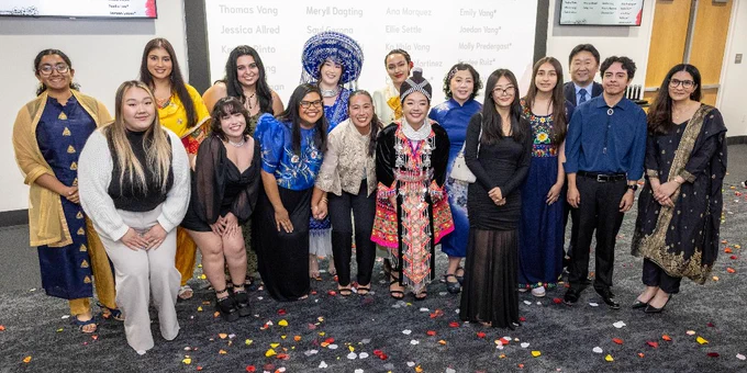 A group of people stand together in a celebratory setting with confetti on the floor. They are dressed in a variety of formal and traditional attire. Behind them, a screen displays names and information labeled "2025 Majors" and "Minors."