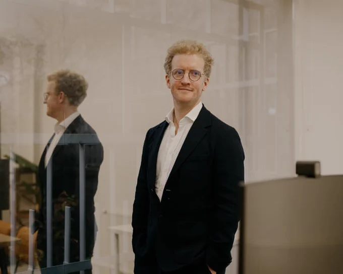 Professional individual in a black suit and white shirt standing confidently in a modern office with glass walls.