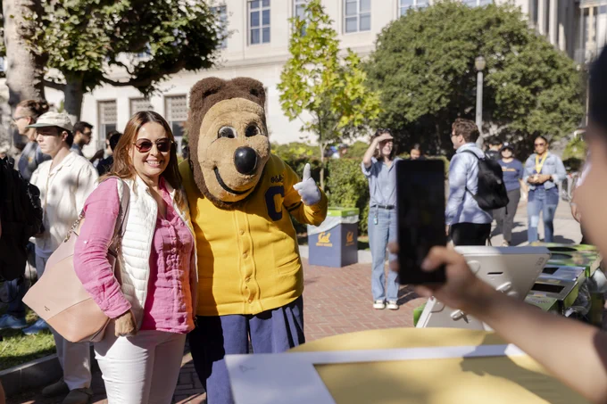 A student poses with the UC Berkeley's mascot, Oski the Bear, in the Campanile Esplanade. Several people are walking and standing in the background, and another person is taking a photo using a smartphone.