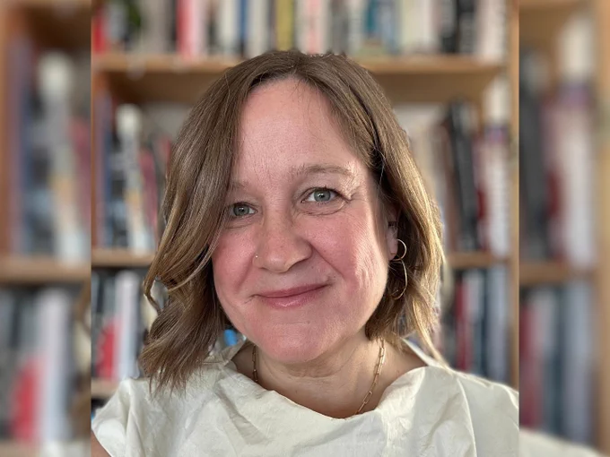 A woman with short blonde hair wearing a white top. A blurred bookcase is in the background.