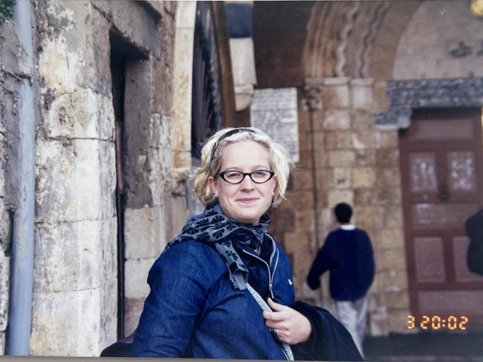 A woman with short blonde hair and glasses wearing a blue jacket and scarf in Lebanon.
