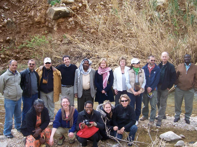 A group of sixteen people posing outdoors in front of a rocky and grassy area. They are standing and crouching on uneven terrain, dressed in casual and outdoor clothing. The setting appears to be a natural environment with sparse vegetation.