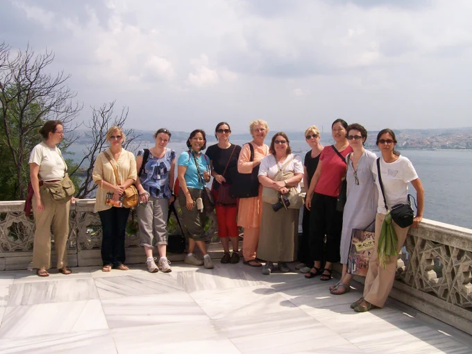 A group of individuals standing together on a marble terrace overlooking the Bosphorus Strait. They are outdoors with a cloudy sky above. Some are holding bags, and there's greenery and distant hills in the background.