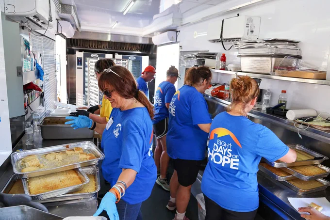 Volunteers wearing blue Eight Days of Hope shirts prepare meals in a kitchen with trays of food on counters and shelves.