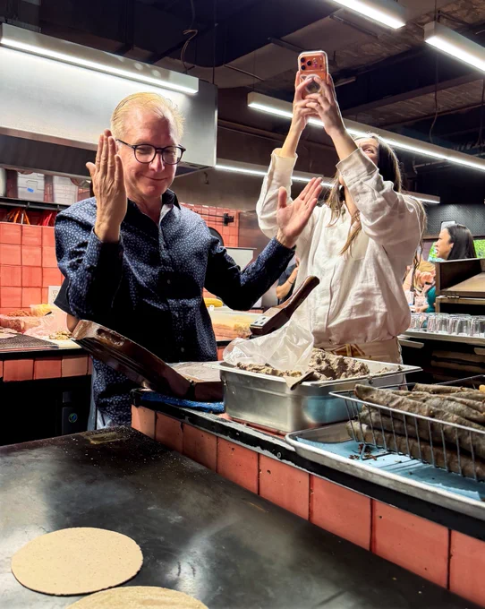 Joz and Ana stand in the kitchen at Maizajo, in front of a flat top stove. Joz raises his hands after he makes a tortilla as Ana takes a photo with her cosmic orange iPhone.