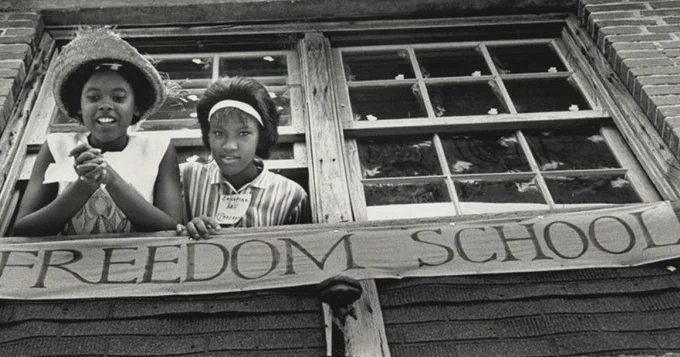 Two young black women poke their heads outside of a window with a banner with the text "Freedom School."