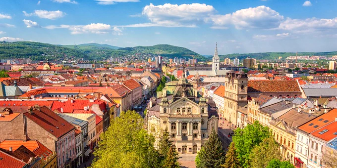 Aerial view of Košice, Slovakia, showcasing colorful historic buildings and streets. The central focus is the ornate State Theater surrounded by lush greenery. Rolling hills and a partly cloudy sky create a picturesque backdrop.
