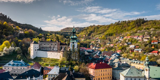 A scenic view of Banská Štiavnica, featuring the historic New Castle and the Church of Saint Catherine amidst colorful rooftops and lush green hills.
