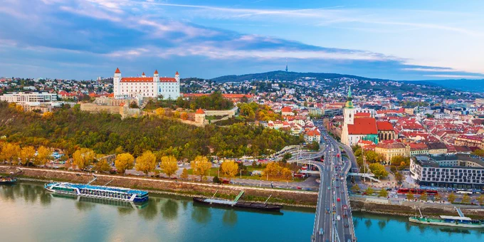 Panoramic view of Bratislava, featuring the historic Bratislava Castle on a hill. The cityscape includes a mix of colorful buildings and the Danube River with a large bridge and boats. The sky is partly cloudy.

