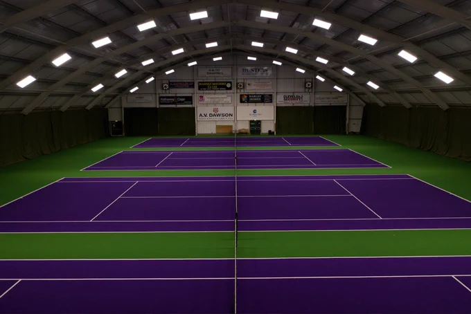 Indoor tennis courts with vibrant purple playing surfaces and green borders under a high arched ceiling with bright lighting.