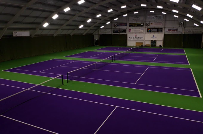 Indoor tennis courts with vibrant purple playing surfaces and green borders under a high arched ceiling with bright lighting.