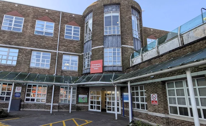 The image shows the exterior of a hospital building, particularly the entrance to the "Emergency Department (A&E)." The building is made of brick with multiple windows, and there are signs indicating a no-smoking area. The entrance has a glass awning and automatic doors.