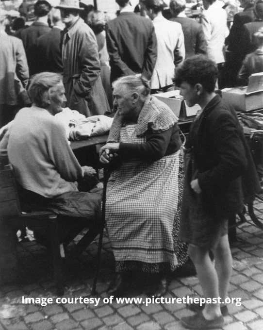 Two elderly women sit facing each other chatting. One woman leans on her walking stick. She wears a long gingham apron and a knitted shawl around her shoulders.  Her hair is in a bun. They sit by a market stall while crowds of shoppers mingle around them. 