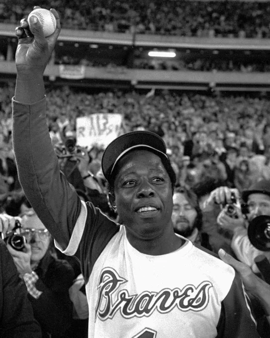 Hank Aaron holds the ball he hit for his 715th career home run in his right hand. He is wearing a white Braves uniform and Braves hat.