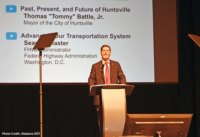 A person stands at a podium on a stage, speaking at an event. Behind them is a large screen displaying topics and speakers. Teleprompters are positioned on either side of the podium.
