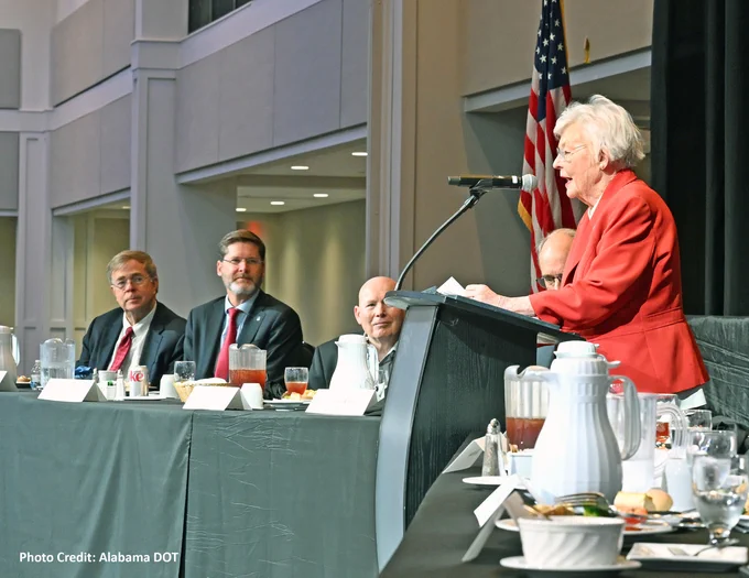 A person speaks at a podium during a formal event with other speakers seated at a long table. An American flag is visible in the background. 
