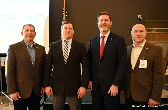 Four smiling men in business suits looking at the camera. US flag is seen in the background.