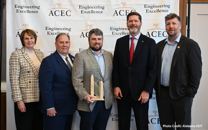 Five individuals in business attire standing facing the camera. Person in the middle holding an award. The backdrop shows ACEC and Engineering Excellence.