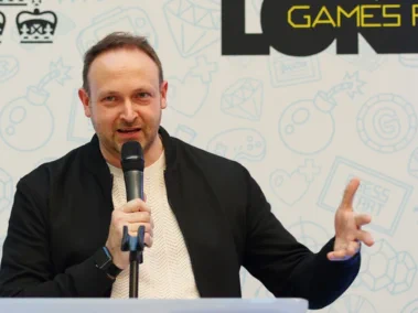 A man with short hair wearing a white jumper and a black jacket. He stands on a stage and holds a microphone in his hand. He is speaking to a live audience.