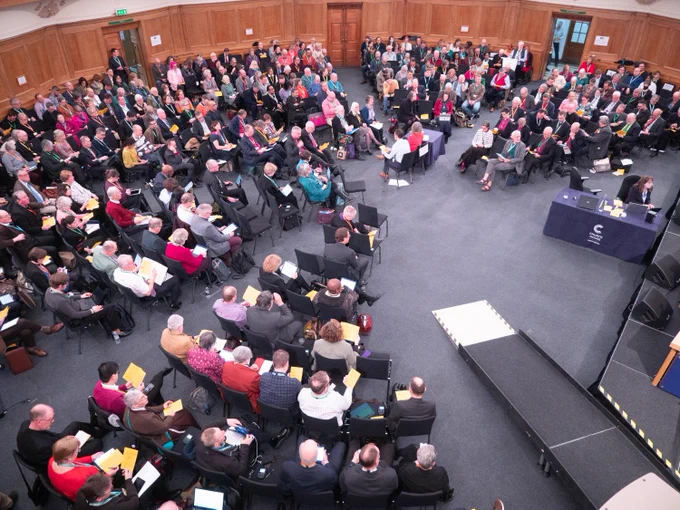 A gathering of Synod members people seated in a circular arrangement in the chamber in Church House, London. At the center, individuals are seated around tables with microphones. A stage banner reads "Church of England." Attendees are holding papers and some appear to be engaged in discussion.