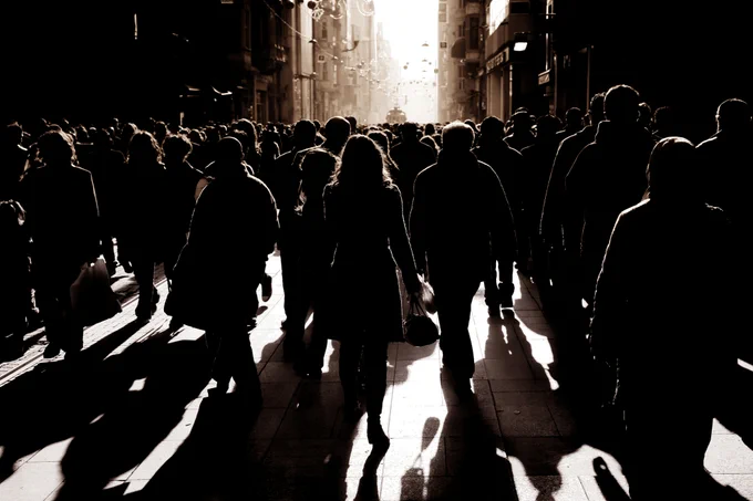 A bustling street scene with many people walking in silhouette, casting long shadows on the ground. The sun sets in the background, creating a warm, glowing sky amidst the urban landscape. Buildings line the street, contributing to the busy city atmosphere.