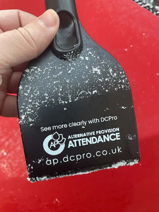 A hand holds a black, branded icescraper which reads "See more clearly with DCPro Alternative Provision Attendance ap.dcpro.co.uk". In the background is a red car bonnet.