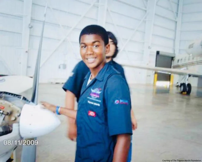 Photo of Trayvon wearing his flight suit with badges running down the front. His hand opposite to the camera is rested on the propeller of a plane. He is smiling at the camera and standing in front of another student in a flight suit who is obscured. A large plane sits in the right corner of the photo.