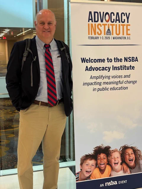 A person in business attire stands next to a sign for the NSBA Advocacy Institute, scheduled for February 1-3, 2026, in Washington, D.C. The sign mentions "Amplifying voices and impacting meaningful change in public education" and shows smiling children.