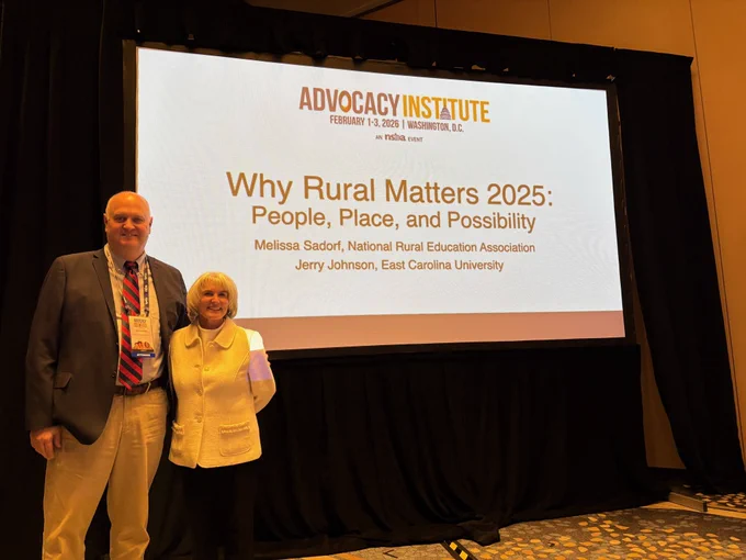 Two individuals stand smiling in front of a screen displaying a presentation titled "Why Rural Matters 2025: People, Place, and Possibility." The screen also lists the names Melissa Sadorf, National Rural Education Association, and Jerry Johnson, East Carolina University. The event is part of the Advocacy Institute in Washington, D.C., dated February 11, 2025.